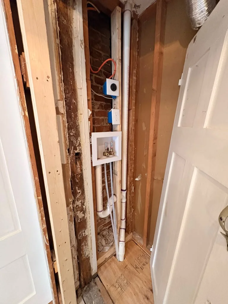 Exposed framing and plumbing in a room under construction, revealing brick wall and floor. by Boston Strong Plastering in Ips